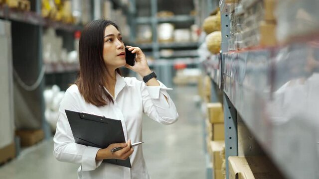 young woman worker talking on a mobile phone and holding clipboard to checking inventory in the warehouse store