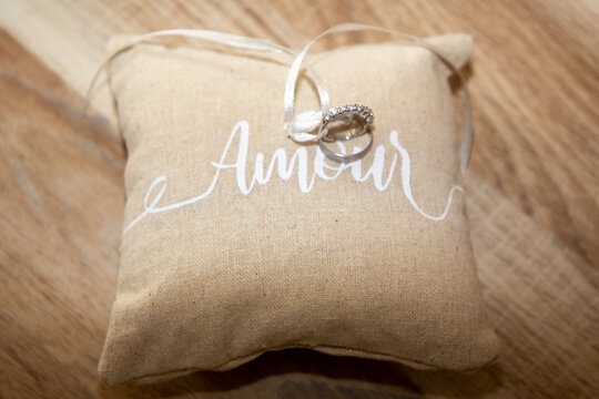 wedding rings on small natural brown pillow with ribbon and amour text means love in french on wooden table background