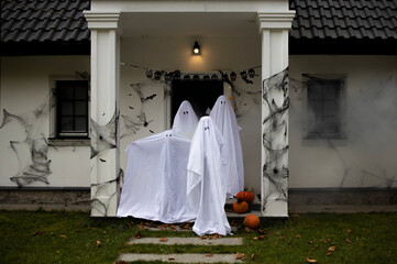 Funny family of four dressed as bedsheet ghosts posing on a porch of their house decorated for a Halloween celebration