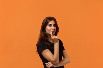 look at me. Portrait of attractive caucasian woman in black t-shirt keeps fore finger near lips. has healthy skin, has beauty smile, looks directly at camera. Isolated over an orange wall