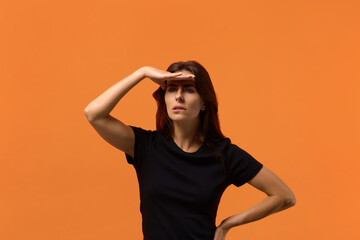 Where is it. Wary caucasian woman in black t-shirt focused into distance, biting lip, searching for something or someone, keeps palm near forehead. Isolated over an orange wall