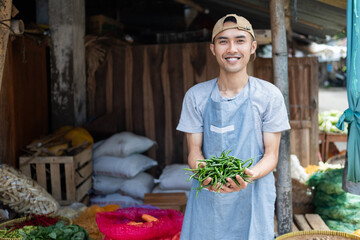 vegetable seller wearing an apron standing holding green chilies in a vegetable stand