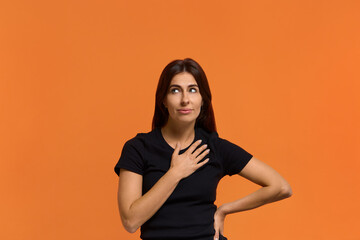 Obraz premium Very unexpected. Portrait of happy caucasian woman in black t-shirt folds arm on chest, expresses gratitude, promises something, has tender smile and long hair. Isolated over an orange wall