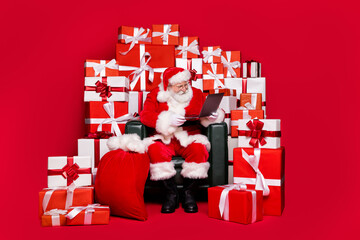 Full length body size view of his he nice handsome bearded focused Santa sit in chair reading e-mail wish list North Pole order delivery isolated bright vivid shine vibrant red color background