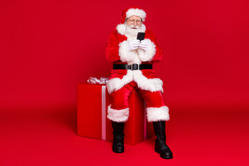 Full length body size view of his he handsome cheerful bearded Santa father wearing warm coat sitting on box use device app delivery celebrate isolated bright vivid shine vibrant red color background