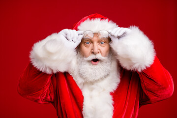 Photo of pensioner old man grey beard excited look hold eyeglasses open mouth shock see female same outfit wear santa costume coat spectacles headwear isolated red color background