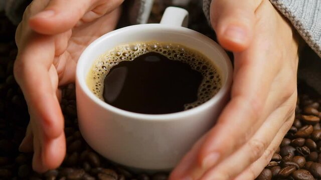 Side view of hands with cup of black coffe on coffee beans background. White cup of black coffee. Delicate woman hands taking small cup of coffee. Selective focus, close up.