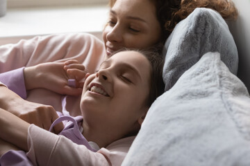 Close up of happy loving Caucasian mother and 13s teen daughter lying relaxing in bed at home together. Smiling caring mom and 12s teenager girl child rest on sofa dream enjoy leisure family weekend.