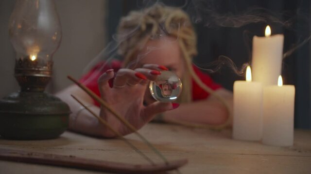 Crystal ball in hands of female medium. Blurry attractive female psychic with dreadlocks scrying future with crystal gazing in rustic house illuminated with candle lights during seance of clairvoyance