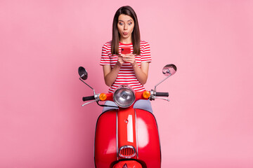 Photo portrait of amazed woman sitting on red moped using smartphone whistling isolated on pastel...