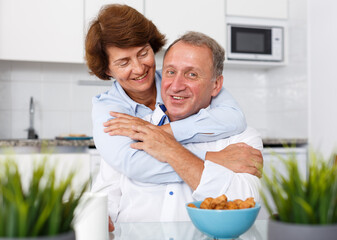 Portrait of mature family couple hugging and sitting at kitchen table while drinking tea