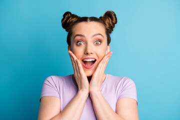Photo of attractive shocked crazy teen lady two funny pretty buns good mood excited expression listen unexpected news arms on cheeks wear casual purple t-shirt isolated blue color background