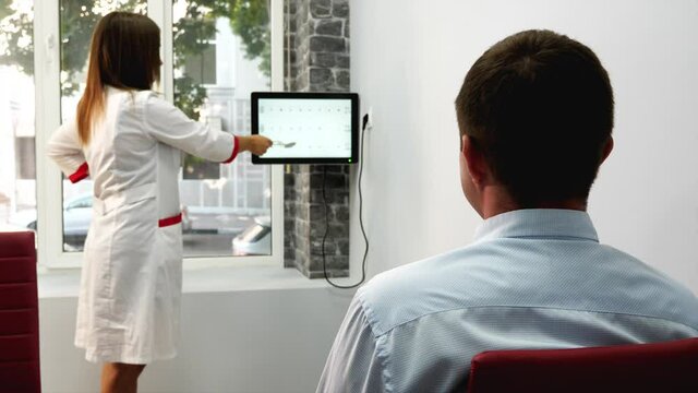 Vision test in optician salon. Man is reads symbols on monitor screen while doctor indicates on characters, rear view. High quality 4k footage