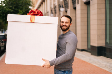 Portrait of smiling delivery man holding large festive box with red bow at outdoors city street, looking at camera. Courier male delivering gift to client. Guy making surprise for girlfriend or wife.