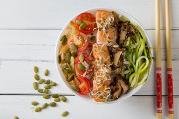 bowl of chinese noodles with salmon shitake mushrooms zucchini onion and tomato on white wooden table