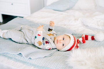 Fototapeta na wymiar the child's first Christmas. a cute little boy in a Santa hat. lying on the bed. playing with a light garland. Christmas concept