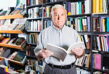 Old czech positive male is choosing book for reading in free time in bookstore.