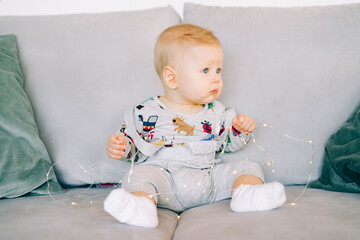 a cute little boy who is teething is sitting in a festive Christmas costume on a gray sofa and playing with a bright light garland.Christmas concept