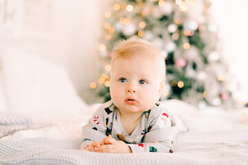 the child's first Christmas. a pensive sad little boy is lying in a festive costume on the bed against the background of a brightly decorated fir tree with a Golden light garland.Christmas concept