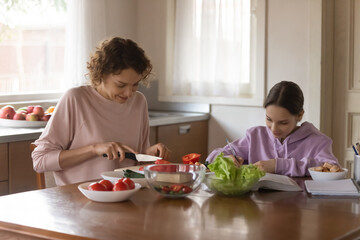 Obraz na płótnie Canvas Happy Caucasian mother sit at table in kitchen cook prepare healthy food for family dinner. Smart teenage daughter schoolgirl study learn prepare homework assignment writing. Daily routine concept.