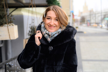 portrait of a beautiful young happy woman in fashionable winter fur coat walking along a city street