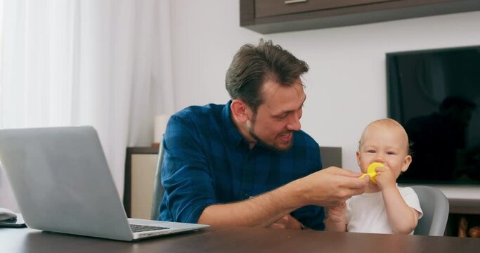 Young bearded man sitting at desk at home with laptop. Cute baby sits at high baby chair, father holds teether which baby bites. Man kisses baby in a cheek. Slow motion
