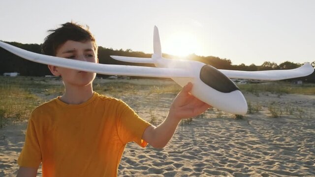Happy boy holding an airplane in his hand is playing and walking along sand on seashore against background of bright summer sun sunset slow motion. Child is playing lifestyle. Travel. Childhood