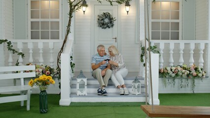 Satisfied senior elderly Caucasian couple sitting and drinking wine in porch stairs at home