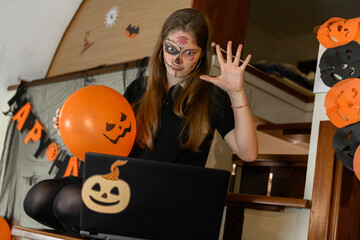 A girl in a Halloween make-up congratulates her friends and family on Halloween with a video call via a laptop. Celebrating at home a distance, online Halloween costume and mask contests