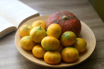 Wooden bowl with tangerines and a pumpkin and open book on a table. Selective focus.