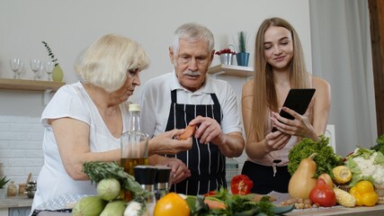 Mature grandparents couple cutting vegetables for salad, listening recipe from girl with tablet