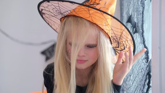 4k. Halloween kids. Portrait blonde girl in witch costume at home. Child puts witch hat on her head. Holiday preparations.