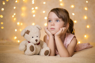 little girl with a toy bear lies on the bed against the background of Christmas lights