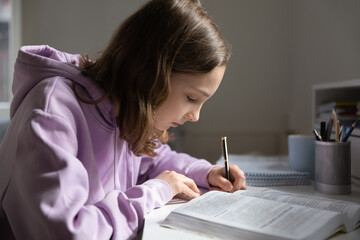Concentrated Caucasian teen girl pupil handwrite in notebook prepare homework assignment at home. Focused smart teenager write read study with textbook at home. Distant education concept.