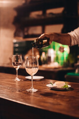 A barman pouring white wine into a glass. Wooden table in a dark bar