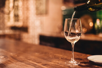 A barman pouring white wine into a glass. Wooden table in a dark bar