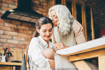 young woman breastfeeds baby and talks to grey haired mother and sitting on wooden chair.