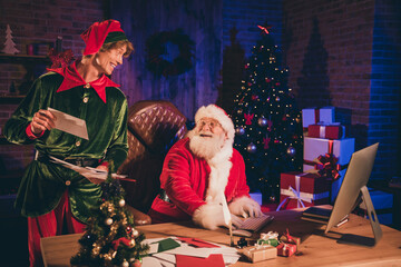 Photo portrait of santa claus and elf looking at each other holding paper mail