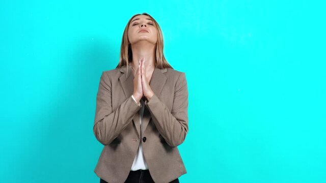 Young woman in business suit holding hands in prayer, asking for help and healing, pleading for forgiveness and support. Studio image indoors, isolated on blue background