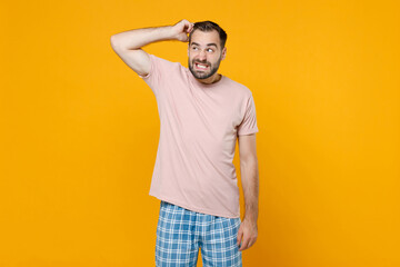 Preoccupied puzzled young bearded man in pajamas home wear put hand on head looking aside while resting at home isolated on bright yellow background studio portrait. Relax good mood lifestyle concept.