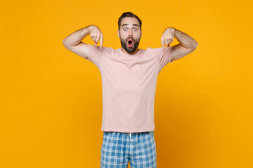 Shocked amazed young bearded man in pajamas home wear point index fingers down on mock up copy space resting at home isolated on yellow background studio portrait. Relax good mood lifestyle concept.