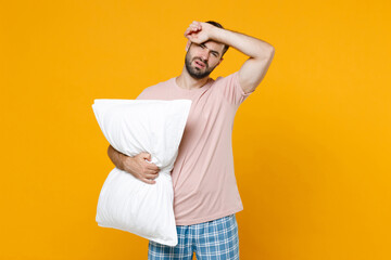 Exhausted young bearded man in pajamas home wear hold pillow put hand on head while resting at home isolated on bright yellow colour background studio portrait. Relax good mood lifestyle concept.