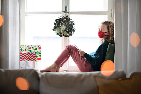Small girl with face mask sitting indoors at home at Christmas, using tablet. Coronavirus concept.
