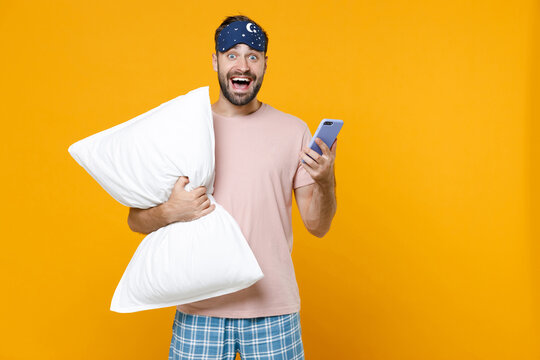 Excited young man in pajamas home wear sleep mask hold pillow using mobile cell phone typing sms message while resting at home isolated on yellow background studio portrait. Relax good mood concept.