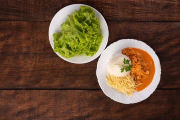 Rice, chicken stroganoff, potato straw and lettuce in white dish, on wooden surface, gray background, top view.