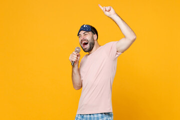 Cheerful young man in pajamas home wear sleep mask sing song in microphone pointing index finger up resting at home isolated on yellow background studio portrait. Relax good mood lifestyle concept.