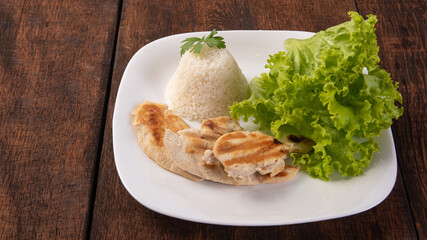 Rice, lettuce and grilled chicken on a white plate, on wooden surface, gray background, selective focus.