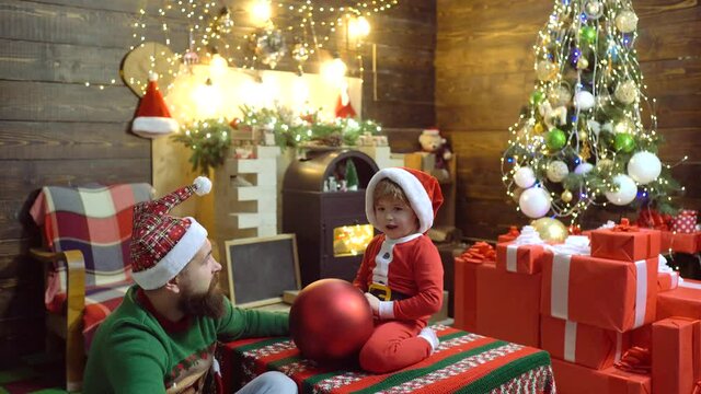 Father and son with bauble at home near Christmas tree and fireplace. Dad and child in Santa hat hold Xmas prasent at home. Kid with parent enjoy New Year.