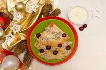 A Christmas dessert on a green and red plate lies on a white wooden table with New Year's decorations. Children's creative food. Pancakes, cupcakes with berries and condensed cream. View from above.