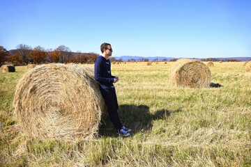 Naklejka na ściany i meble Young brunette man in warm jacket leaning against haystack in field with bales of hay. he looking to the right. Primorsky kray, Russia. Beauty in nature. Copy space